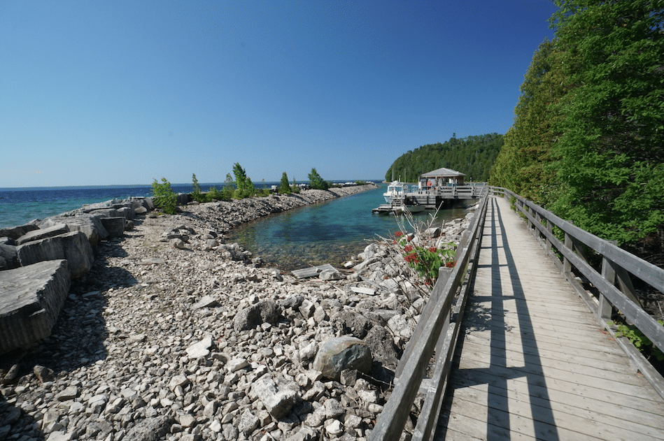 A wooden boardwalk runs alongside a rocky shoreline leading to a small dock with boats, set against clear blue skies and tranquil turquoise waters.