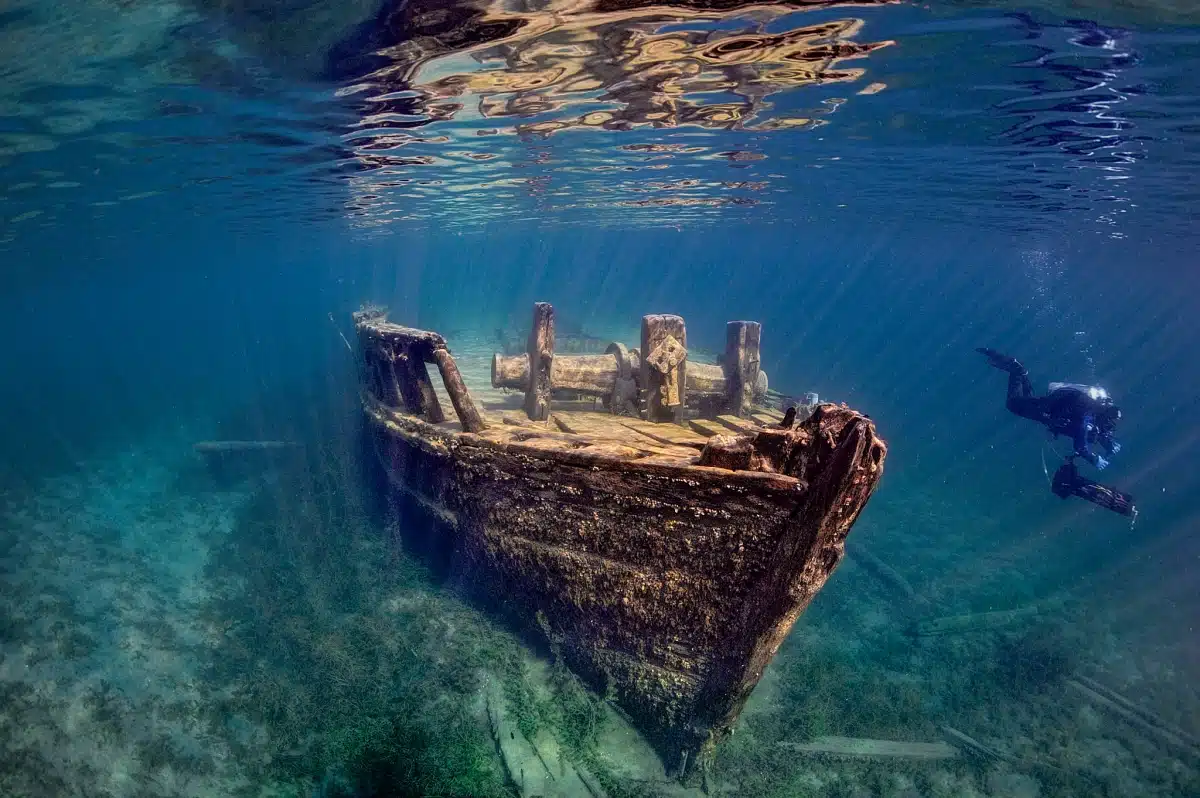 An underwater image showing a sunken ship with textures of decay and a person diving near it, highlighting the eerie tranquility of the aquatic environment.