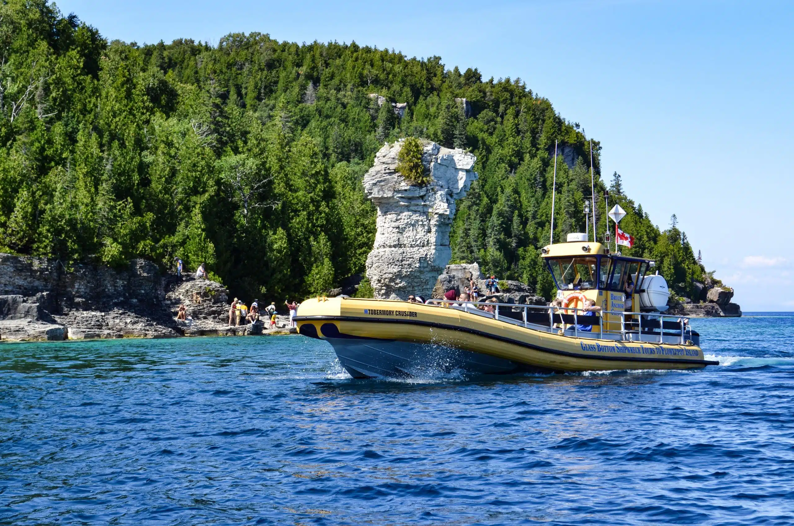 A yellow tour boat is moving across clear blue water near a rocky shore with green trees. Unique rock formations are visible in the background.