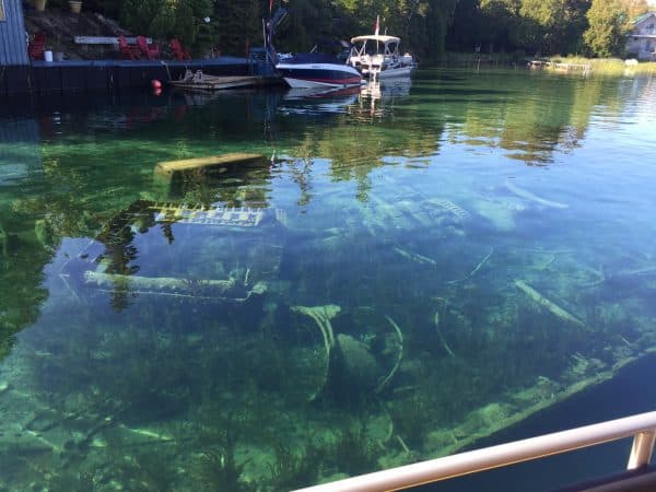 The image shows crystal clear water providing a view of submerged trees, with boats docked nearby and a glimpse of shoreline structures in the background.