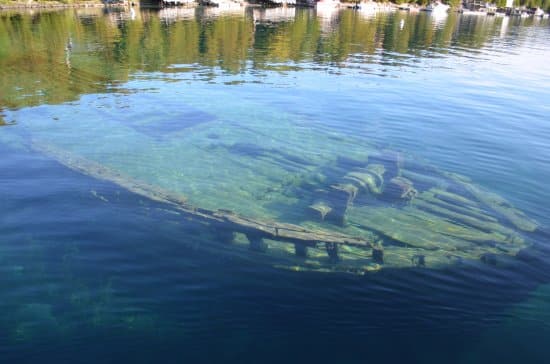 The image shows a submerged shipwreck visible through clear blue water, with trees and reflections on the water's serene surface.