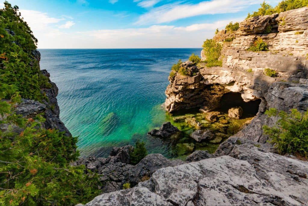 A serene coastal landscape featuring rugged cliffs, a turquoise sea, green vegetation, and a grotto, under a partly cloudy blue sky.