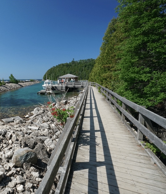 A wooden boardwalk runs alongside a rocky shoreline leading to a small dock with boats, set against clear blue skies and tranquil turquoise waters.