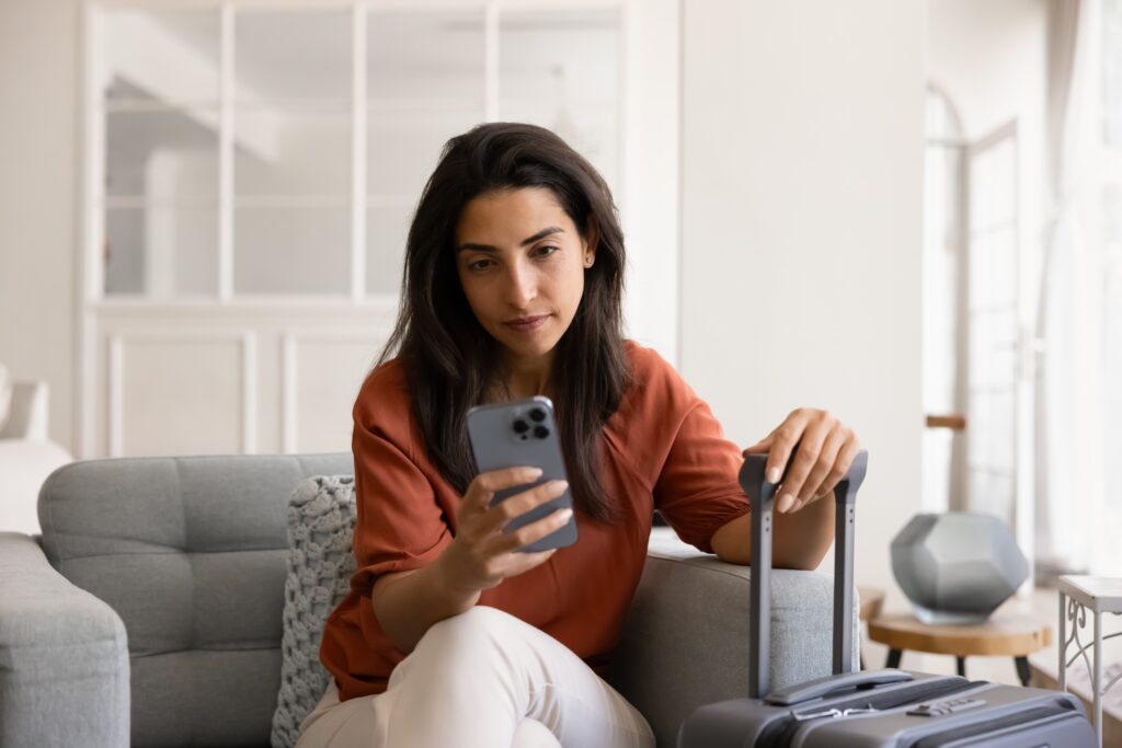 Woman in an apartment holding her phone in one hand and suitcase handle in the other searching for travel accommodations before leaving home